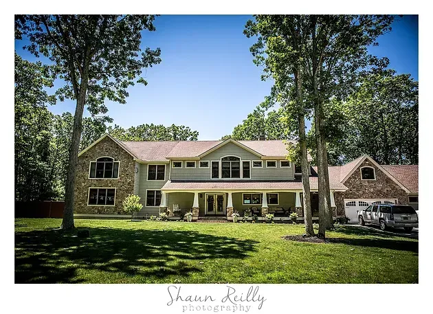 Large two-story house with beige siding, brick accents, and trees in a sunny yard