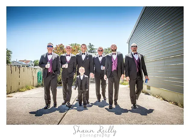 Six people in formal suits posing outdoors on a bright driveway under a blue sky.