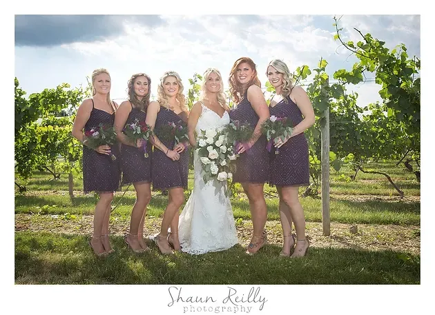 Bride with five bridesmaids in dark dresses posing outdoors in a vineyard.