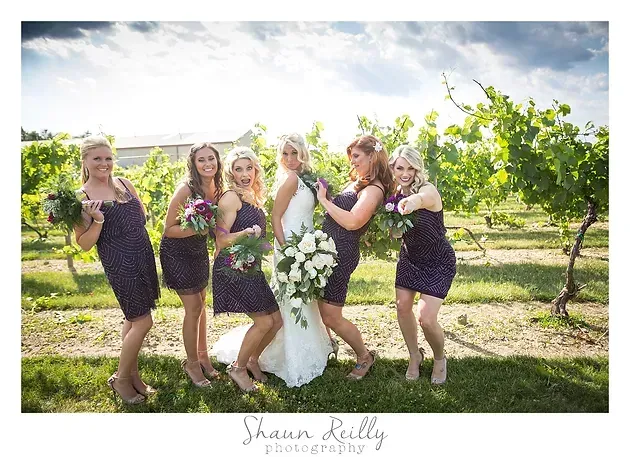 Bridesmaids pose with a bride in a vineyard, holding bouquets and smiling in matching dark dresses.