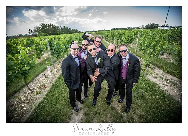 Seven men in suits posing together in a vineyard, smiling toward the camera.