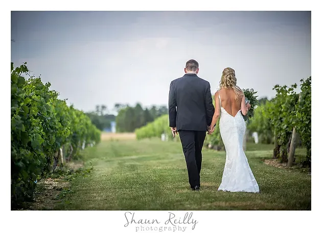 Couple in formal attire walking away down a vineyard path, bride in a white gown and groom in a black suit.