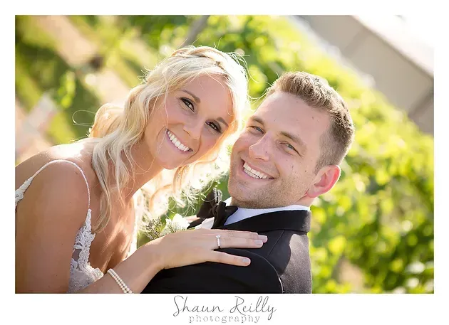 Smiling couple in wedding attire posing outdoors with greenery in the background