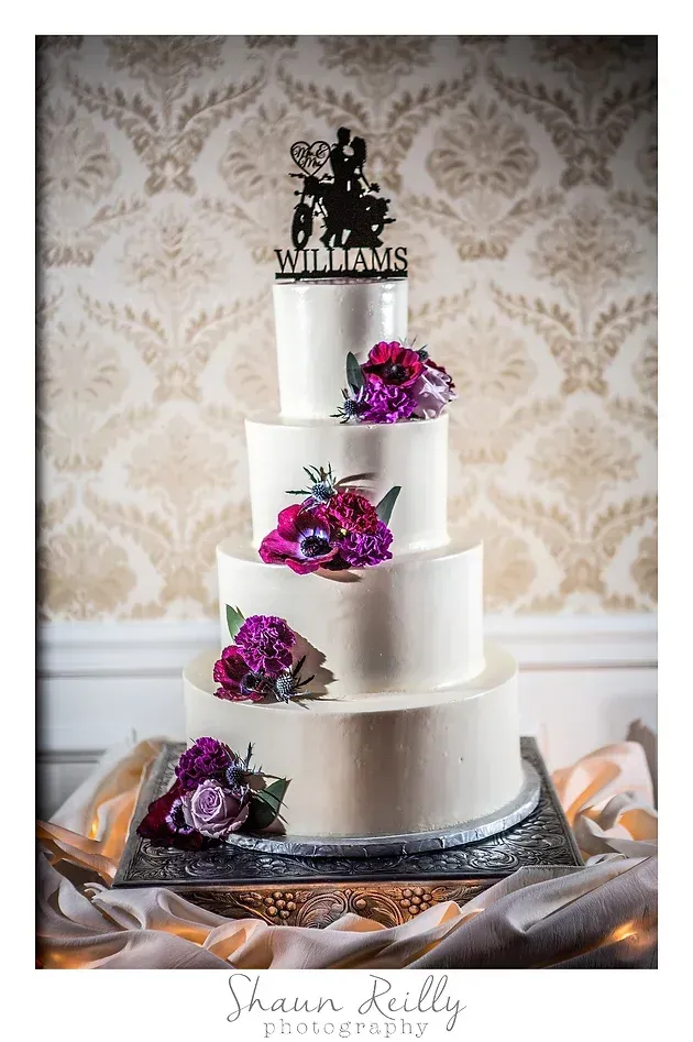 Four-tier white wedding cake with purple flowers and a black topper on a silver stand