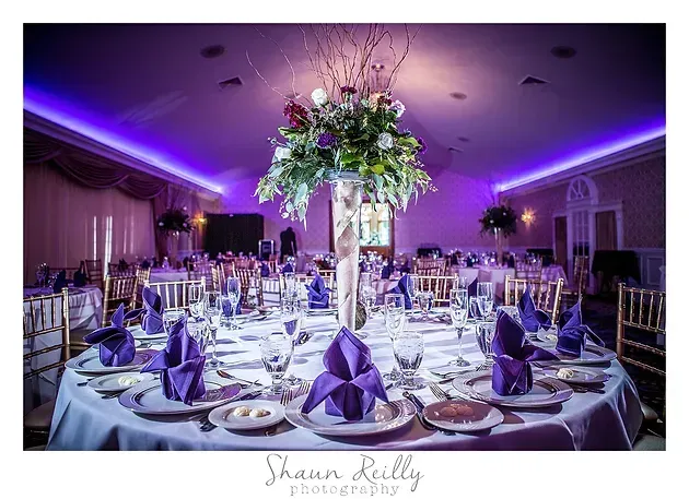 Elegant purple-lit wedding reception with round tables, white linens, and a tall floral centerpiece