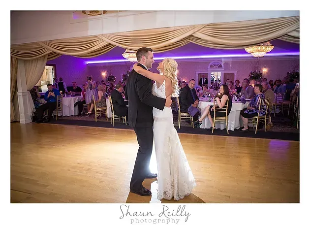 Bride and groom slow dancing at a wedding reception with purple lighting and seated guests in the background