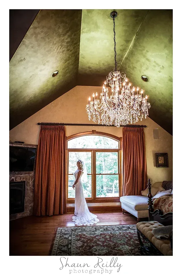 Bride in white gown standing by a large window in a rustic bedroom with chandelier and wood bed