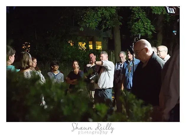 People gathered outdoors at night, listening to a speaker in a dimly lit garden.