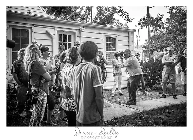 People gathered outdoors near a house while one person speaks and gestures.