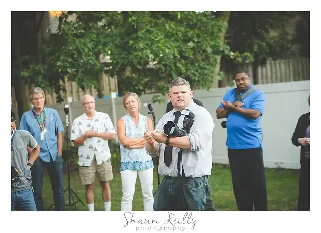Man speaking to a small group outdoors near a white fence, with several people listening behind him.