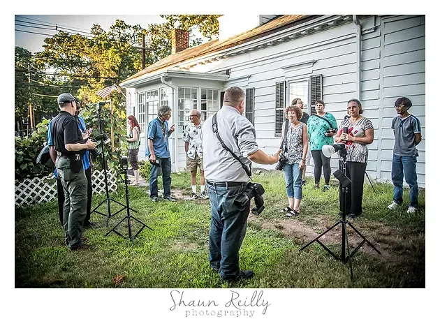 Group of people standing in a yard beside a white house, chatting around black tripod stands