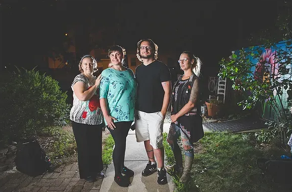 Four people standing together outdoors at night, smiling in a garden path with colorful lights.