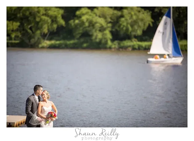 Couple in formal wear by a lake, with a sailboat in the background.