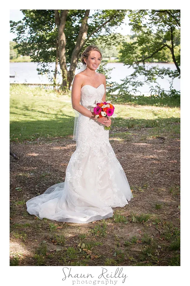 Bride in a white lace gown holding a colorful bouquet outdoors by a lakeside tree.