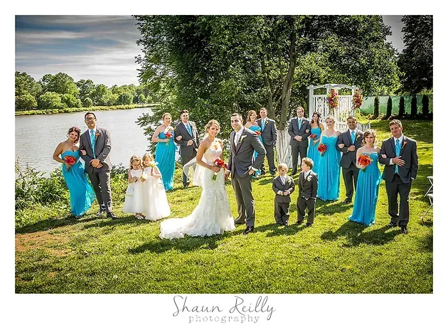 Wedding party posing by a lake, with bride and groom at center under trees.