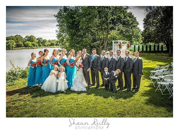 Wedding party posing outdoors by a lake, with bridesmaids in turquoise and groomsmen in gray suits.