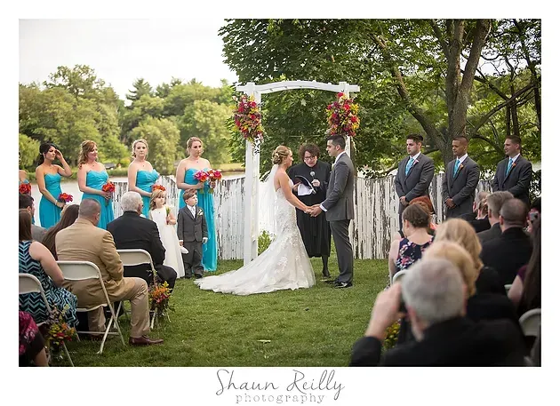 Outdoor wedding ceremony under a white arch, bride and groom kissing as guests watch on lawn