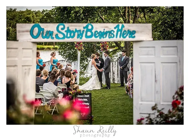 Wedding ceremony under a white arch with “Our Story Begins Here” sign, couple and guests outdoors