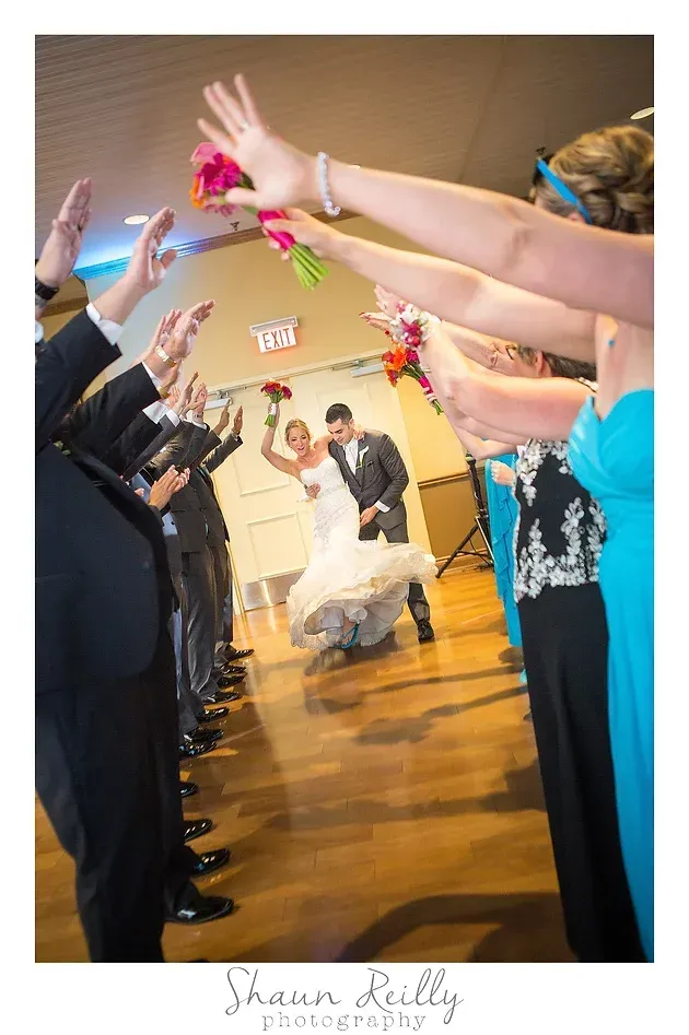 Bride and groom walk under raised arms as guests toss flower petals in a reception hall
