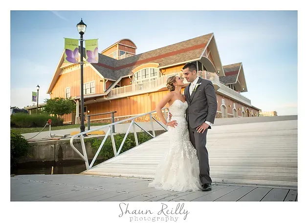 Bride and groom pose in wedding attire beside a rustic building and bridge under a bright sky
