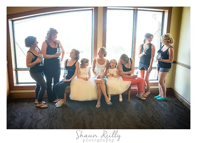 Six women in dresses and formal wear chat and pose by large windows in a bright room.