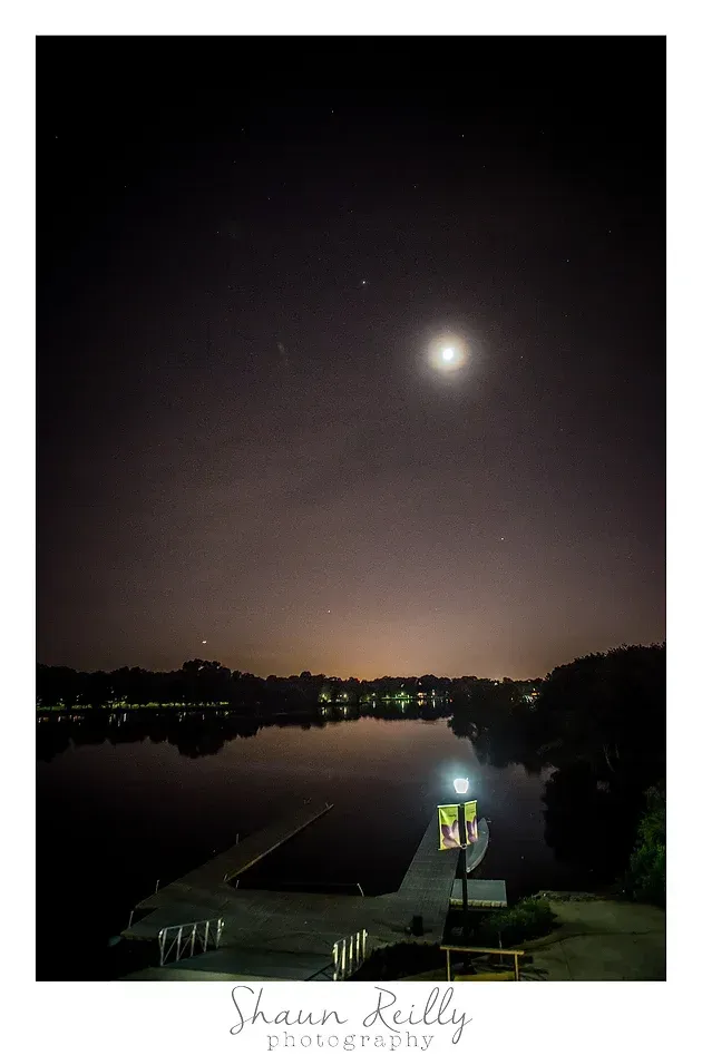 Moonlit river at night with a bright moon, distant city lights, and a lit dock in the foreground