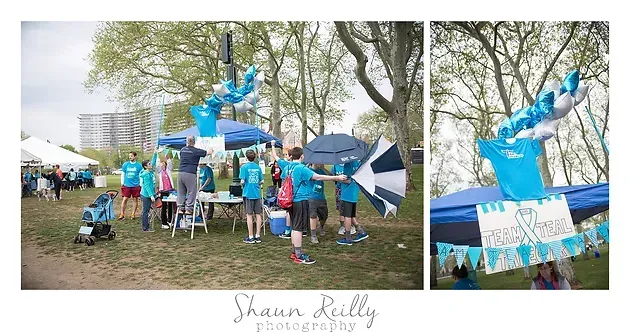 People gathered under trees around a blue inflatable structure at an outdoor event with tents and banners.