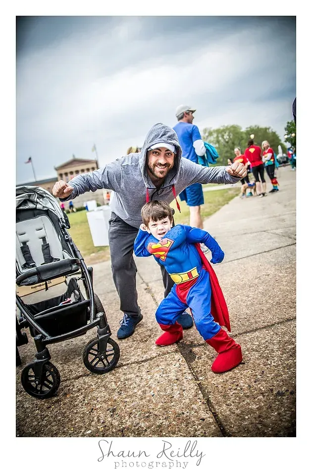 Man in gray and child in blue superhero costume posing playfully outdoors on a cloudy day