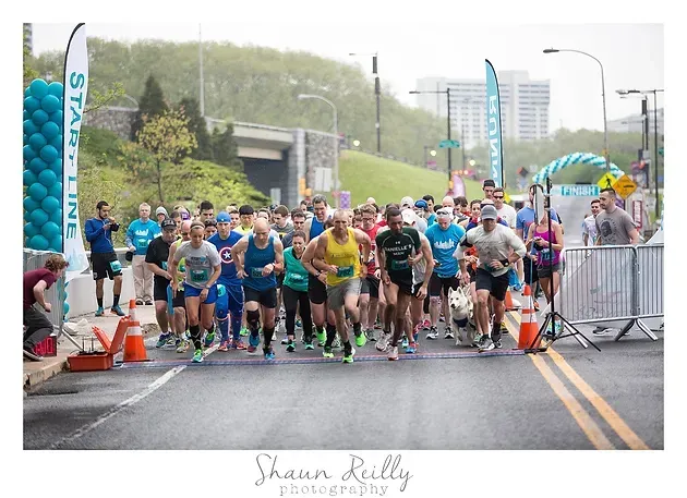 Runners gathered at the starting line of a road race in light rain, with barriers and banners lining the street.