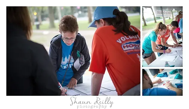 Volunteers helping people sign up at a table, with forms and pens under a white canopy