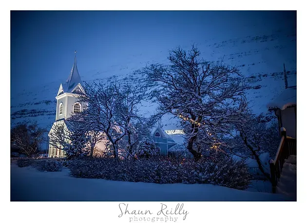 Snowy church steeple at dusk, framed by bare trees in a blue winter landscape.