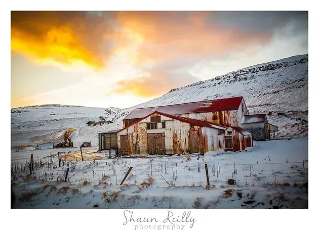 Snow-covered farmhouse and barn at sunset, with warm orange clouds over a rural hillside.