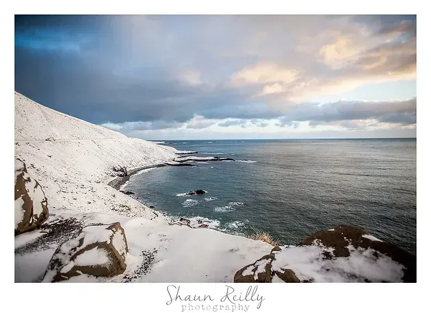 Snowy white coastline meeting a calm gray-blue sea under a cloudy sunset sky