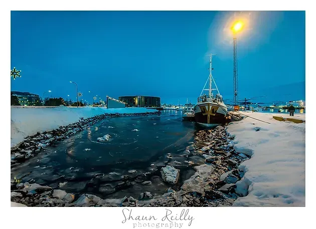 Snowy harbor at dusk with a docked boat, icy water channel, and glowing streetlamp