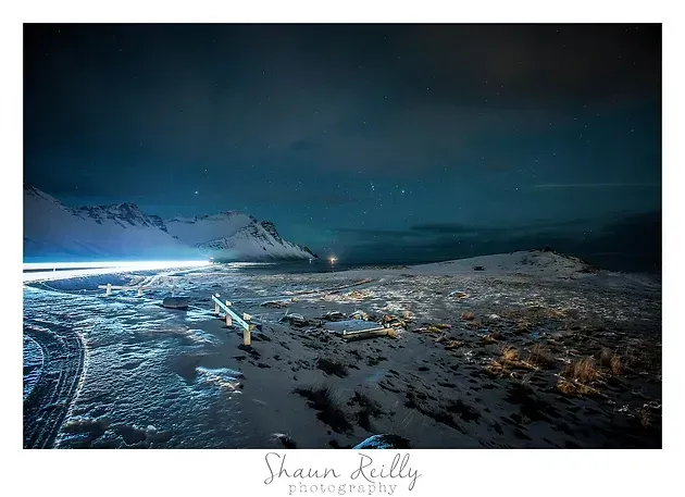 Snowy beach at night with icy shoreline, dark sea, and mountains under a blue sky.
