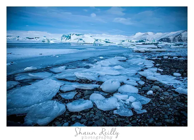 Frozen shoreline with cracked blue ice and distant snowy mountains under a cloudy sky
