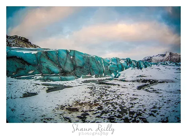 Turquoise glacier under a pastel sky, with snow-covered ground and dark rocky patches.