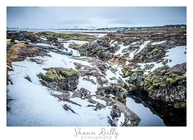 Snow-covered rocky stream winding through a rugged coastal landscape under a cloudy sky