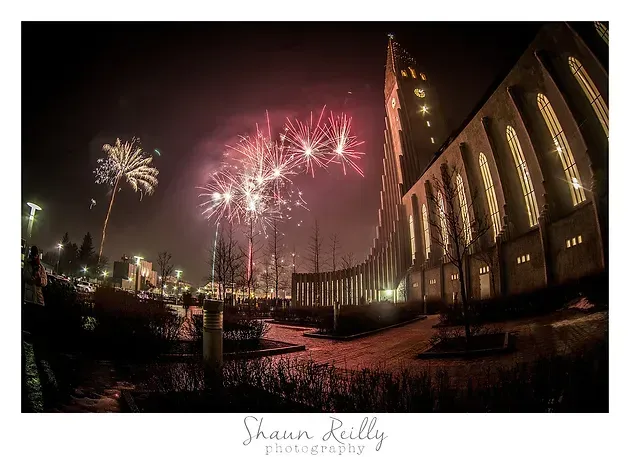 Fireworks bursting over a lit cathedral beside a river at night