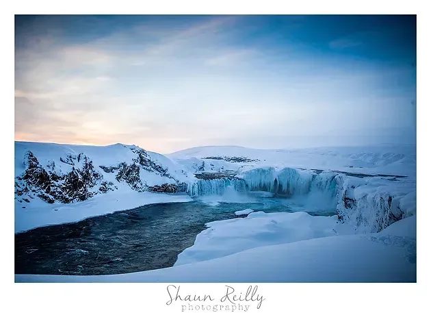 Snowy coastal landscape with icy cliffs, a dark river, and a pale blue sunrise sky