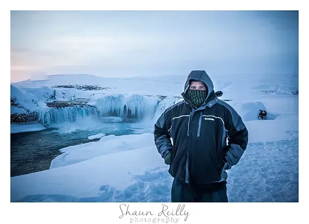 Bundled person stands on icy shore before a frozen waterfall under a pale blue sky