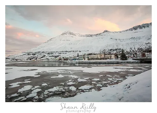 Snowy mountain overlooking a frozen lake at sunrise, with reflections and a small town below