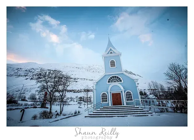 Snow-covered white church with a red door under a blue winter sky, surrounded by snowy hills and bare trees