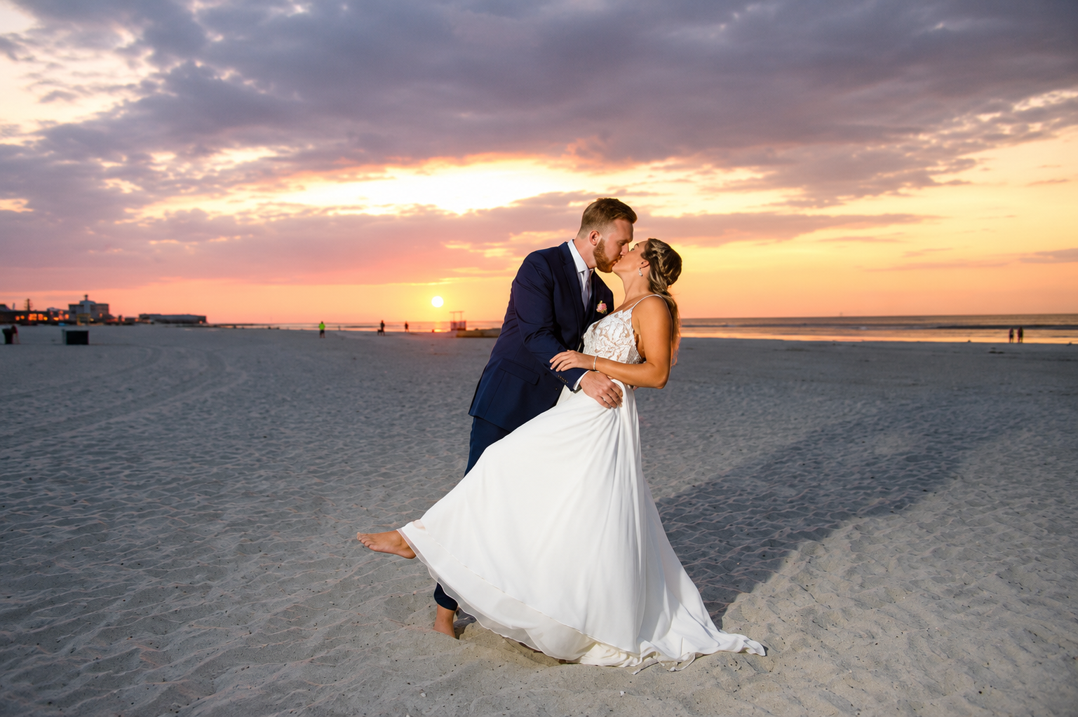 Bride and groom kissing on a beach at sunset, bride in white gown, ocean and pastel sky behind them
