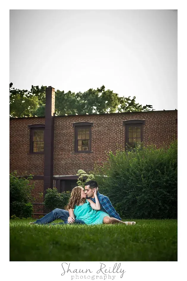 Couple sitting on grass in front of a brick building, with trees and a cloudy sky behind them.