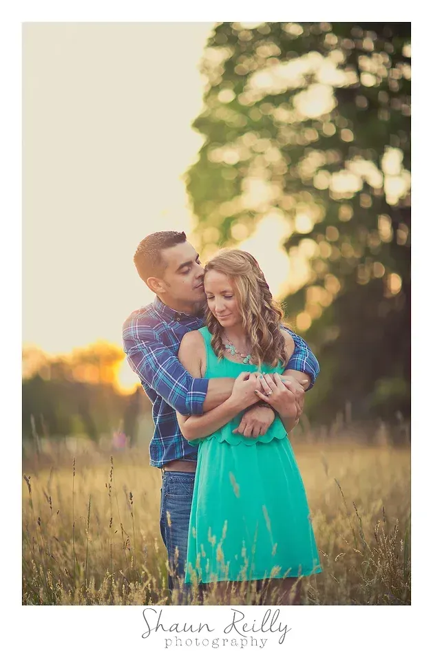 Couple embracing in a sunlit field at sunset, with tall grass and trees in the background.