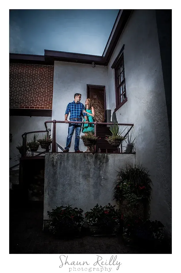 Two people standing on a balcony outside a white building, with a cloudy sky above.
