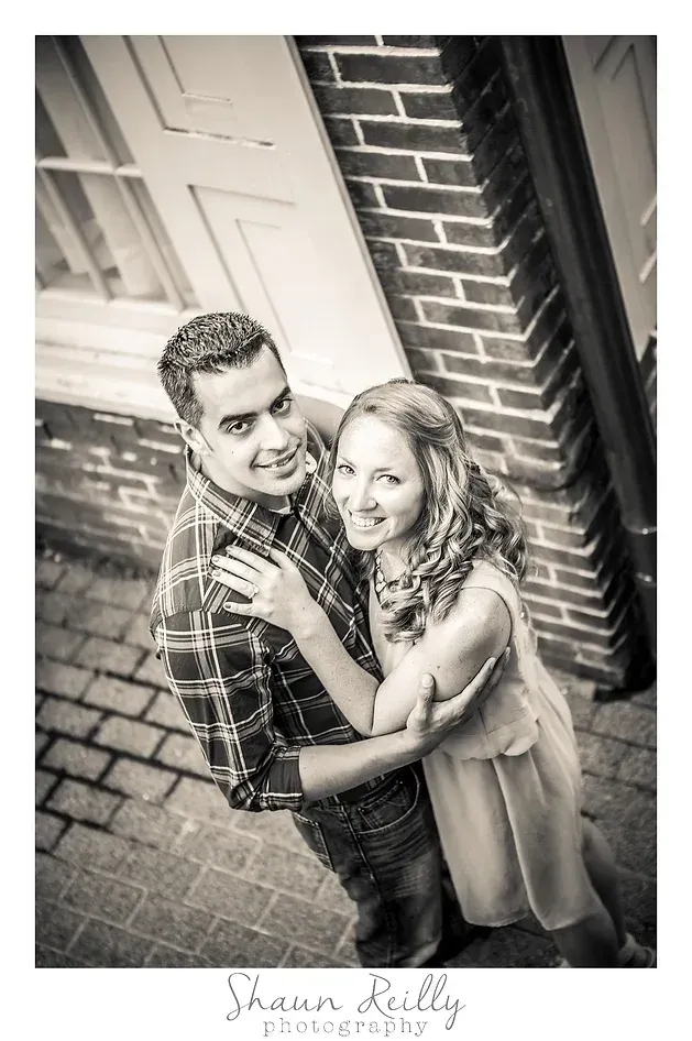 Couple embracing and smiling on a brick walkway beside a wall, in a black-and-white portrait.