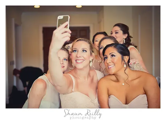 Five women taking a selfie indoors, smiling and dressed in formal attire