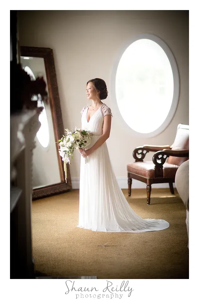 Bride in a white gown holding a bouquet beside a large oval window in a softly lit room